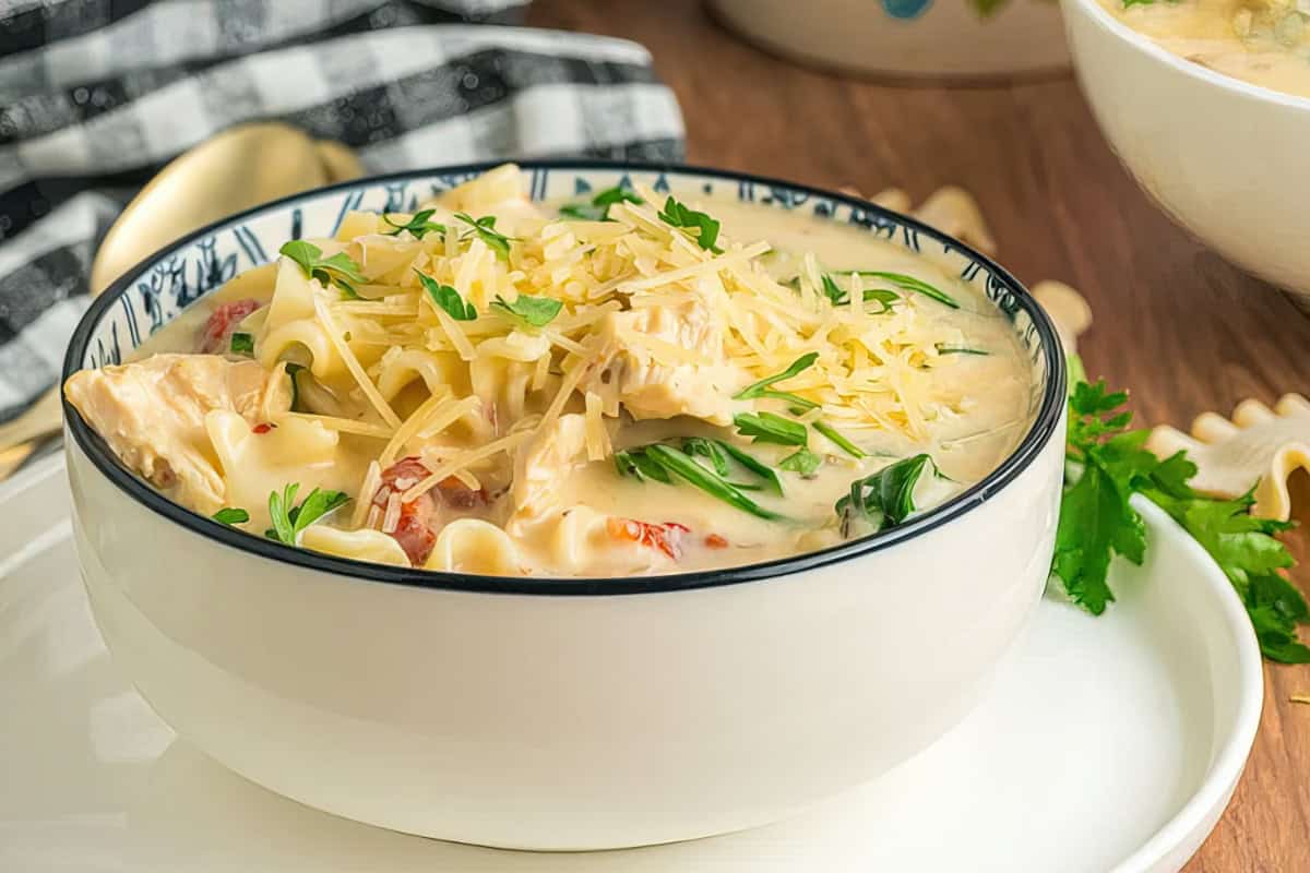 A creamy pasta dish in a white bowl topped with grated cheese and garnished with chopped parsley. The pasta is in a thick sauce with visible spinach and sun-dried tomatoes, set on a wooden table next to a black and white checkered napkin.