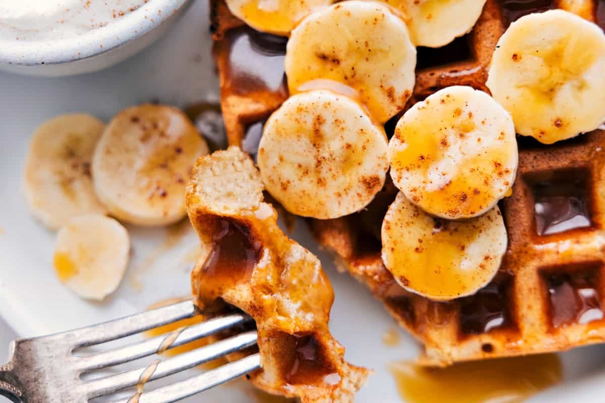 A close-up of waffles topped with banana slices and drizzled with syrup. A fork holds a bite-sized piece with syrup. Extra banana slices are on the side. The waffles have a crispy texture and the syrup glistens in the light.