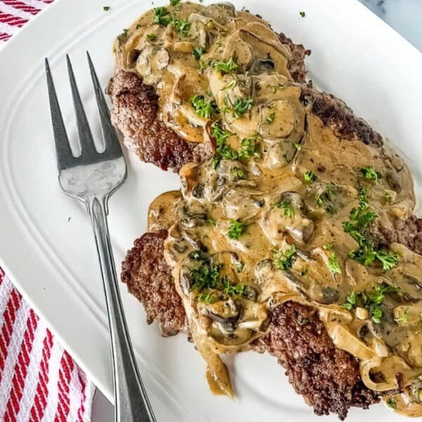 Two pieces of cooked steak topped with creamy mushroom sauce and chopped parsley on a white plate with a serving fork.