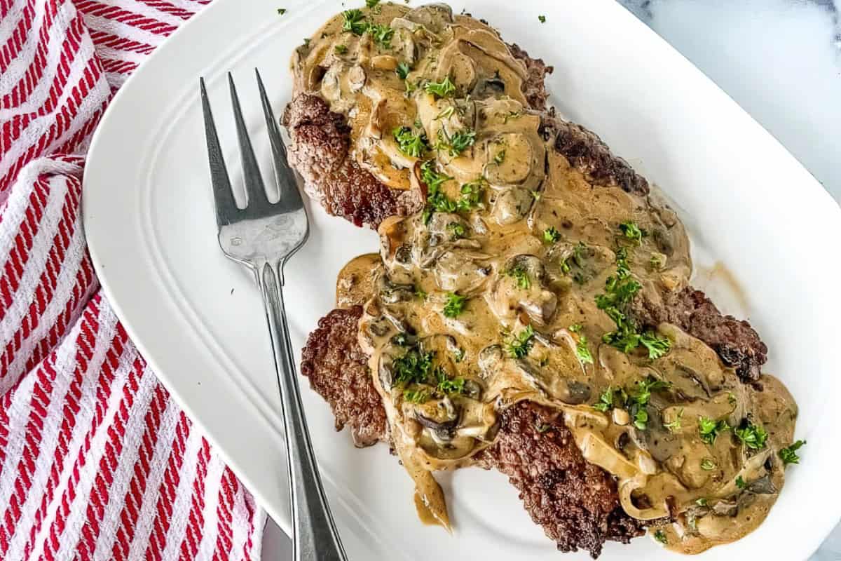 A delicious plate of Salisbury steak topped with creamy mushroom gravy, garnished with chopped parsley. A silver fork rests on the white oval plate beside a red and white striped cloth.
