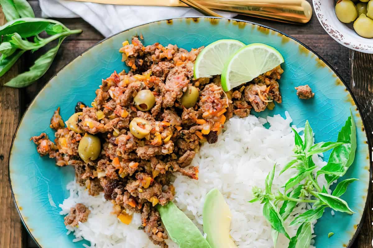 A vibrant plate featuring seasoned ground beef with olives and spices, served with white rice. Garnished with lime slices, a sprig of green herbs, and an avocado slice, displayed on a turquoise plate.
