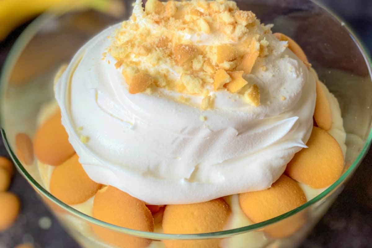 A glass bowl filled with banana pudding. The pudding is topped with a layer of whipped cream, crumbled cookies, and surrounded by whole vanilla wafers.
