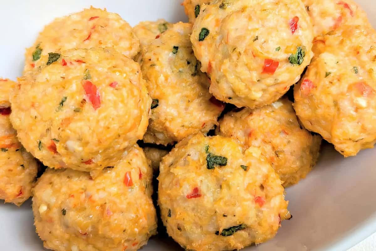 A bowl of golden-brown chicken meatballs mixed with red bell pepper and green herbs. The meatballs have a slightly textured surface and are closely packed together.
