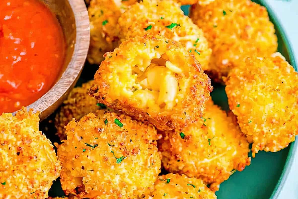 Close-up of crispy macaroni and cheese balls on a green plate with a wooden bowl of red dipping sauce. One ball is cut open, revealing the creamy mac and cheese filling. Garnished with herbs.