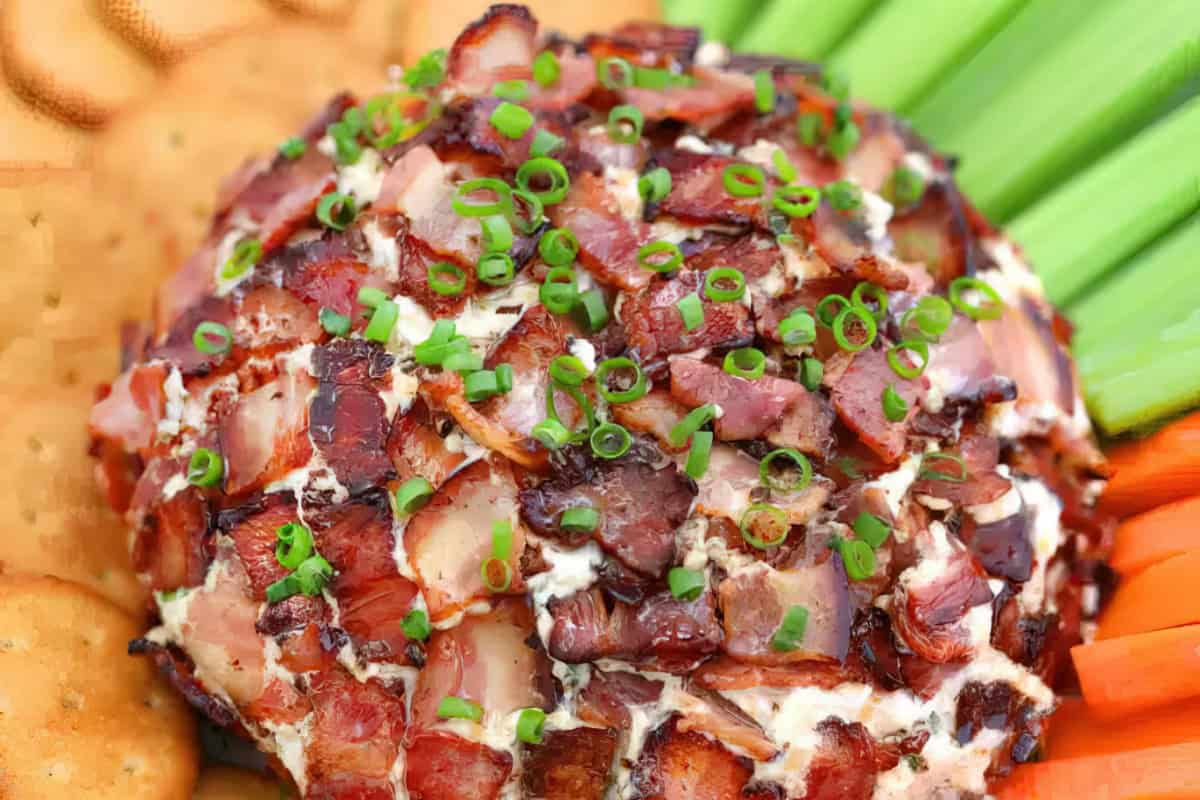 A bacon-covered cheese ball garnished with chopped green onions. It is surrounded by celery and carrot sticks on one side and round crackers on the other.