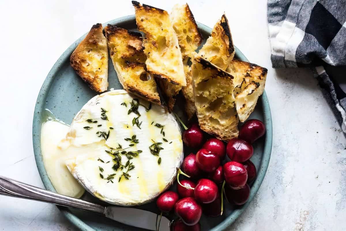 A plate with melted cheese topped with herbs, surrounded by toasted bread and red cherries. A knife rests on the plate, and a checkered napkin is in the top right corner.