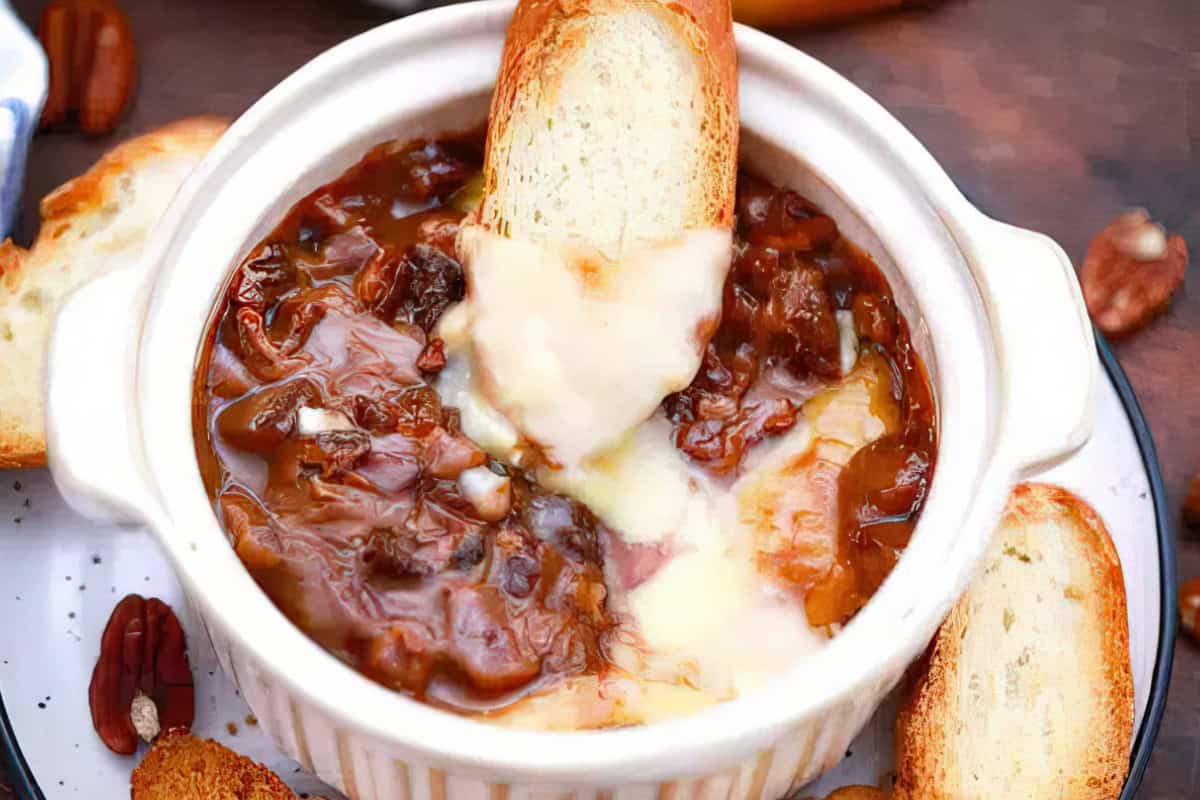 A white bowl filled with French onion soup topped with melted cheese and caramelized onions. A slice of toasted bread is partially dipped in the soup. Slices of bread and pecans are scattered around the bowl.