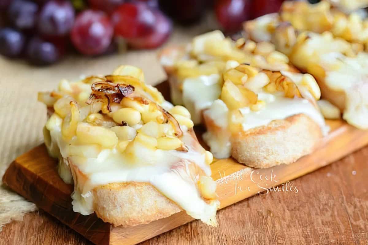 Toasted bread topped with melted cheese, diced apples, walnuts, and caramelized onions on a wooden board. Red grapes are blurred in the background.