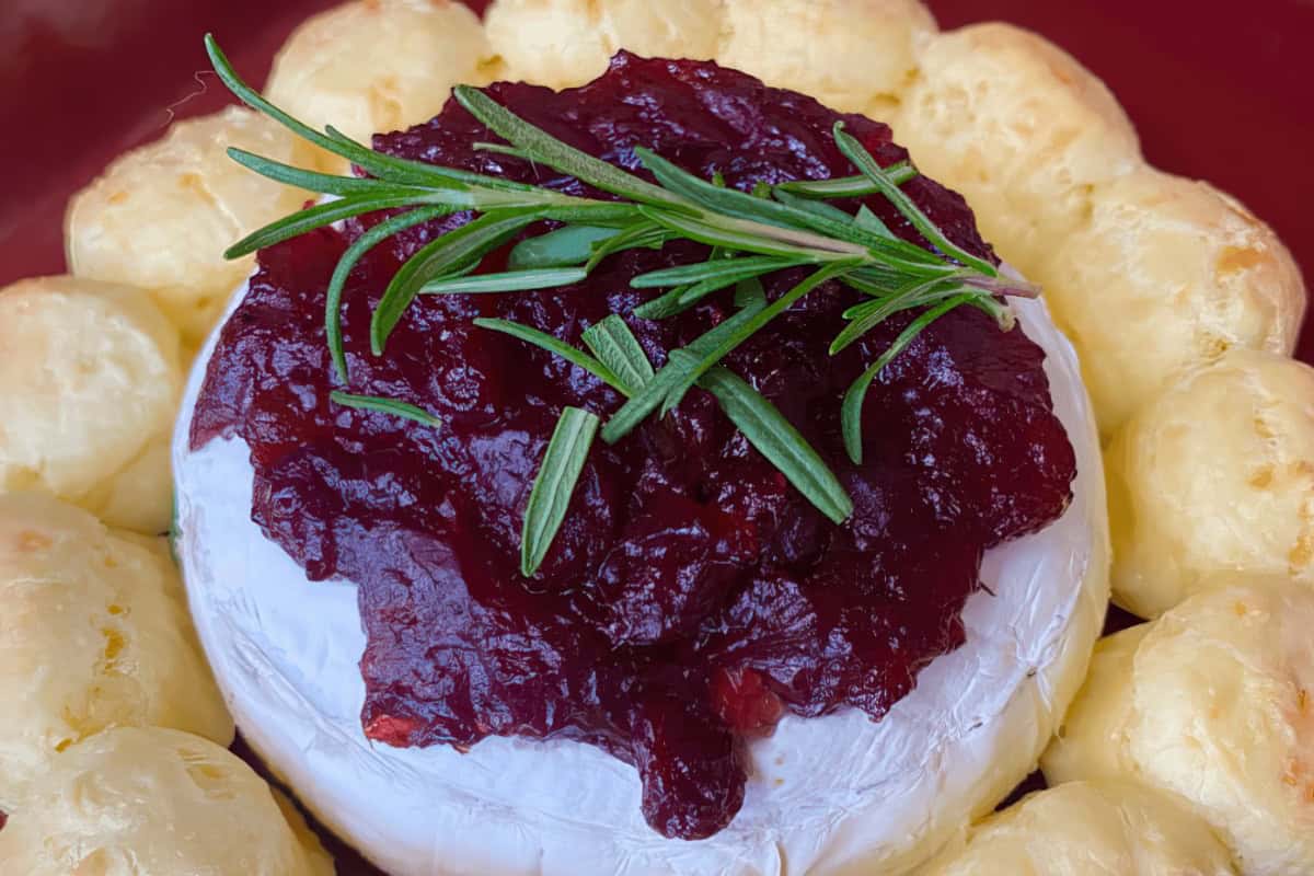 A round of baked brie cheese topped with a cranberry sauce and garnished with fresh rosemary, surrounded by a ring of puff pastry.