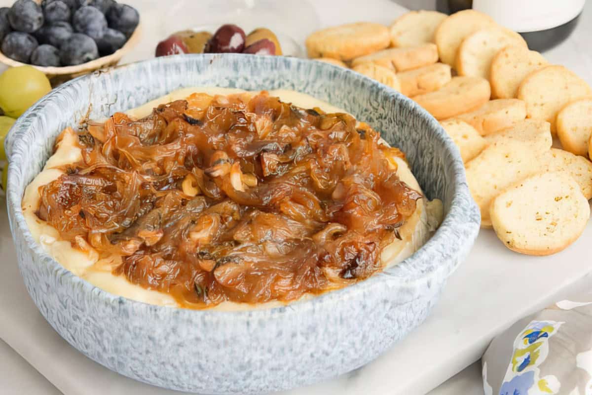 A ceramic bowl filled with caramelized onions on top of creamy melted cheese, surrounded by slices of toasted baguette. A bowl of blueberries and grapes is visible in the background.
