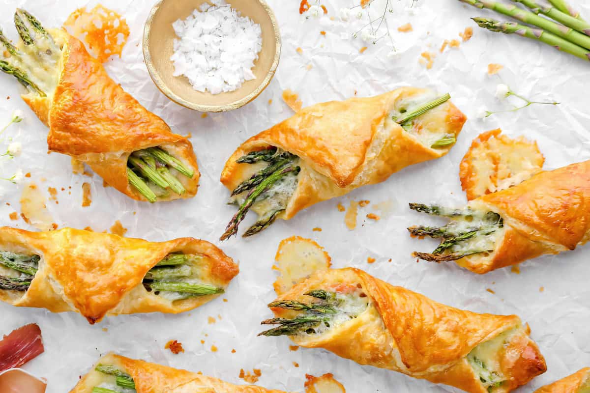 The image depicts golden brown puff pastry wraps containing asparagus spears and melted cheese, placed on parchment paper. A small bowl of salt is positioned in the center.