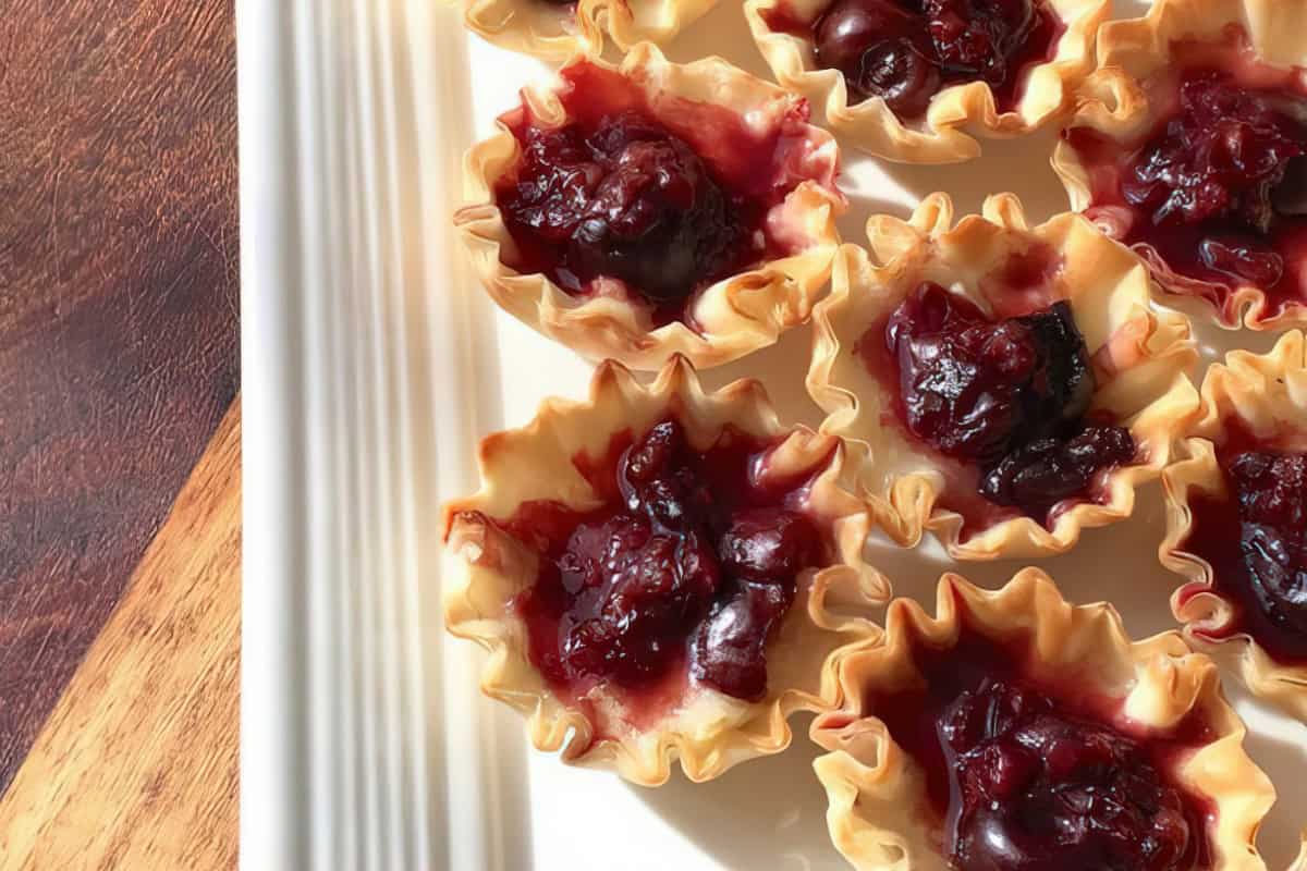 Mini phyllo cups with cherry filling are arranged on a white plate, with part of a wooden surface visible.