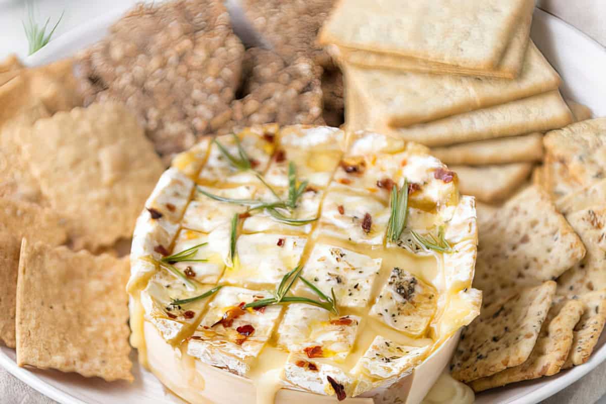 The image shows a platter featuring baked Brie cheese adorned with herbs and accompanied by an assortment of crackers.