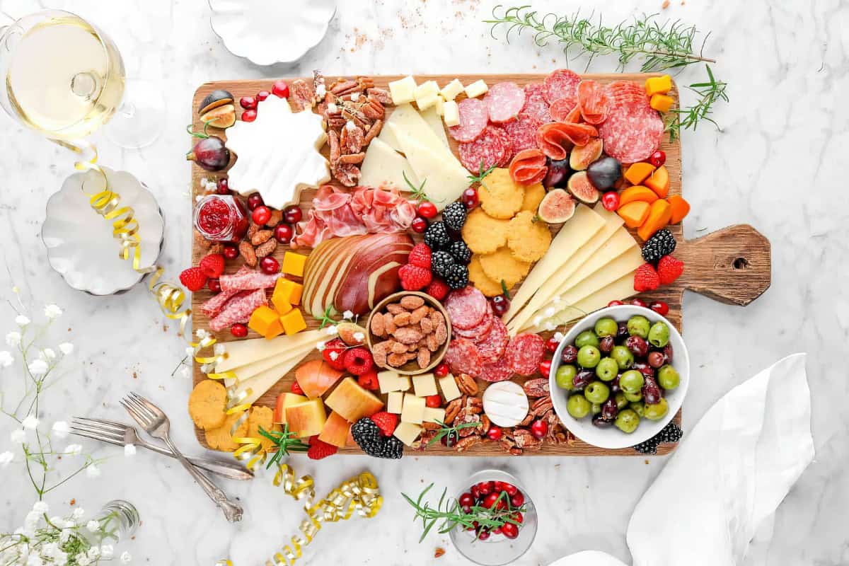 A charcuterie board with cheeses, meats, fruits, nuts, and crackers is set on a marble surface. Nearby are a bowl of grapes and a glass of white wine.