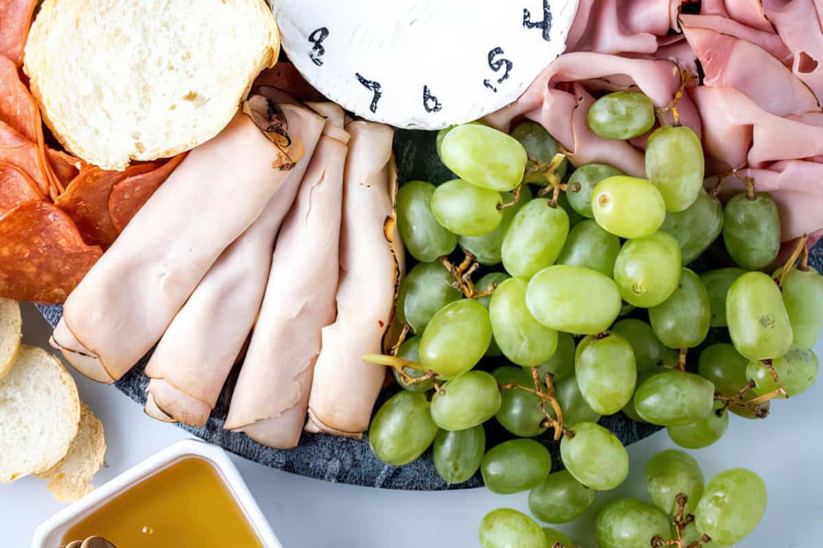 The image displays a charcuterie board featuring sliced meats, a round cheese wheel, green grapes, sliced bread, and a small dish of honey.