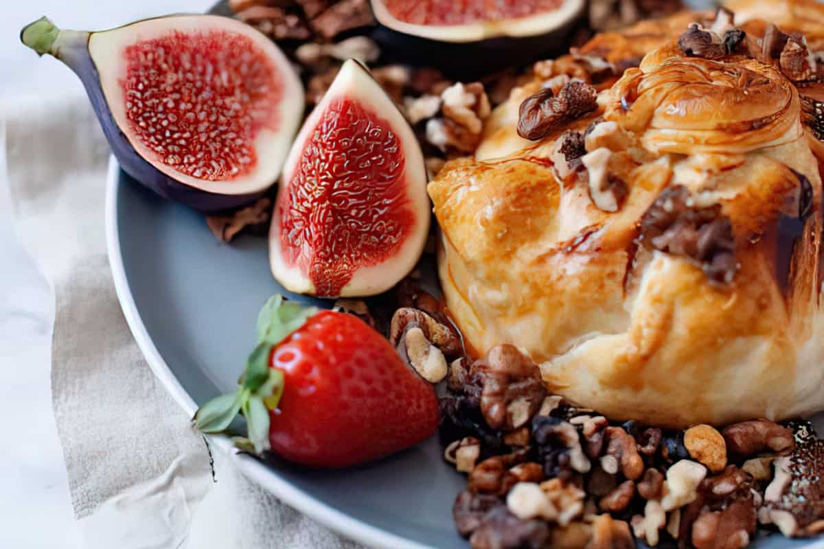 The image shows a plate featuring baked brie topped with nuts, surrounded by fresh figs and a strawberry.