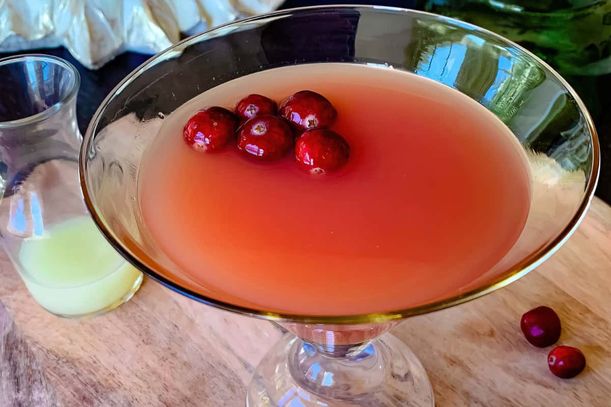 A martini glass filled with a pink cocktail, garnished with several red cranberries. A small glass pitcher with a pale yellow liquid is next to the cocktail, all set on a wooden surface.
