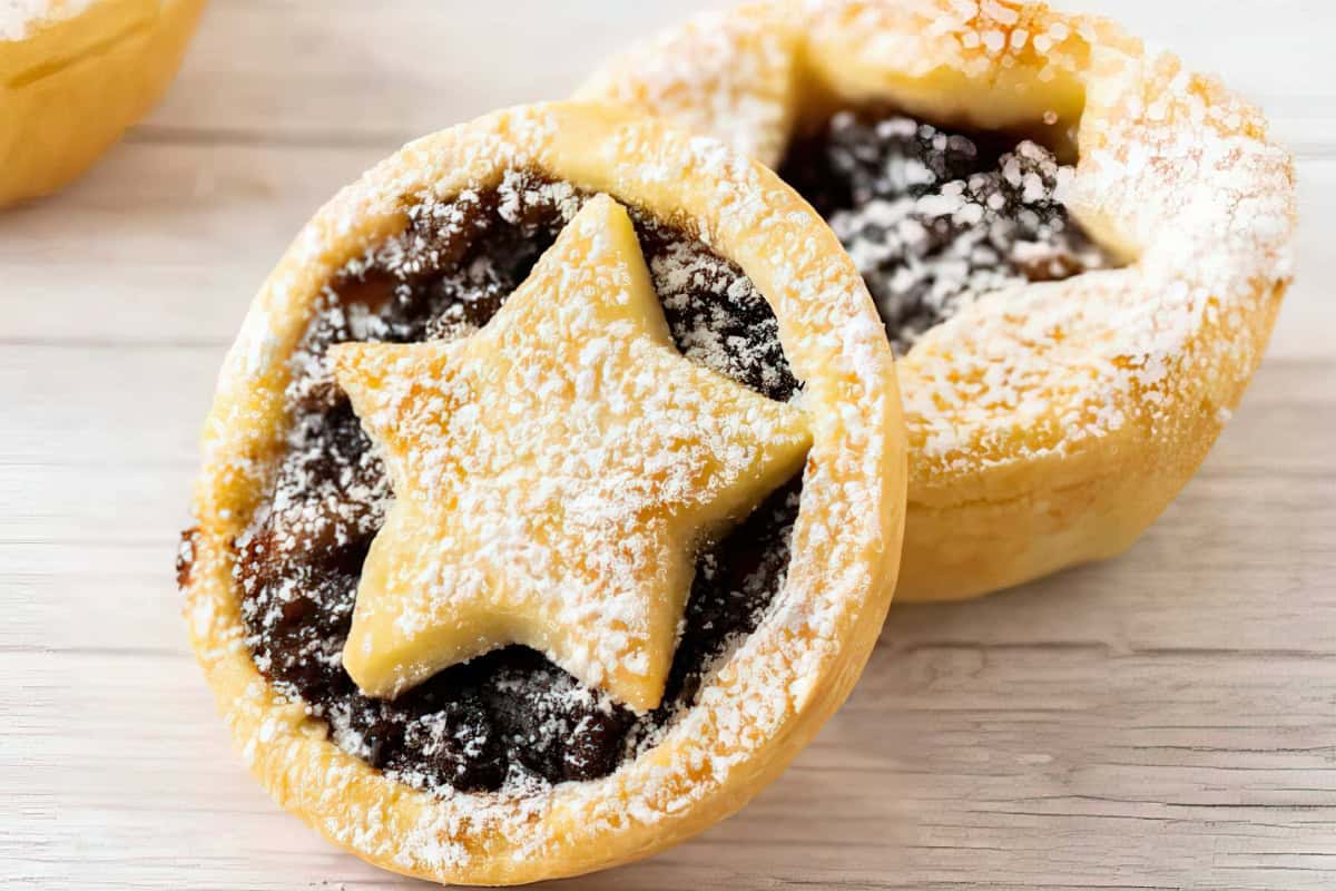 Two baked mince pies with star-shaped pastry tops, dusted with powdered sugar, on a light wooden surface.