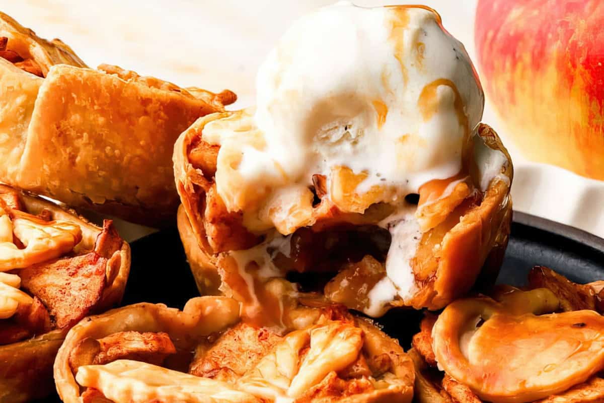 Mini apple pies topped with melting vanilla ice cream on a black plate. The pies are filled with caramelized apple chunks and have decorative crusts. A fresh apple is visible in the background.