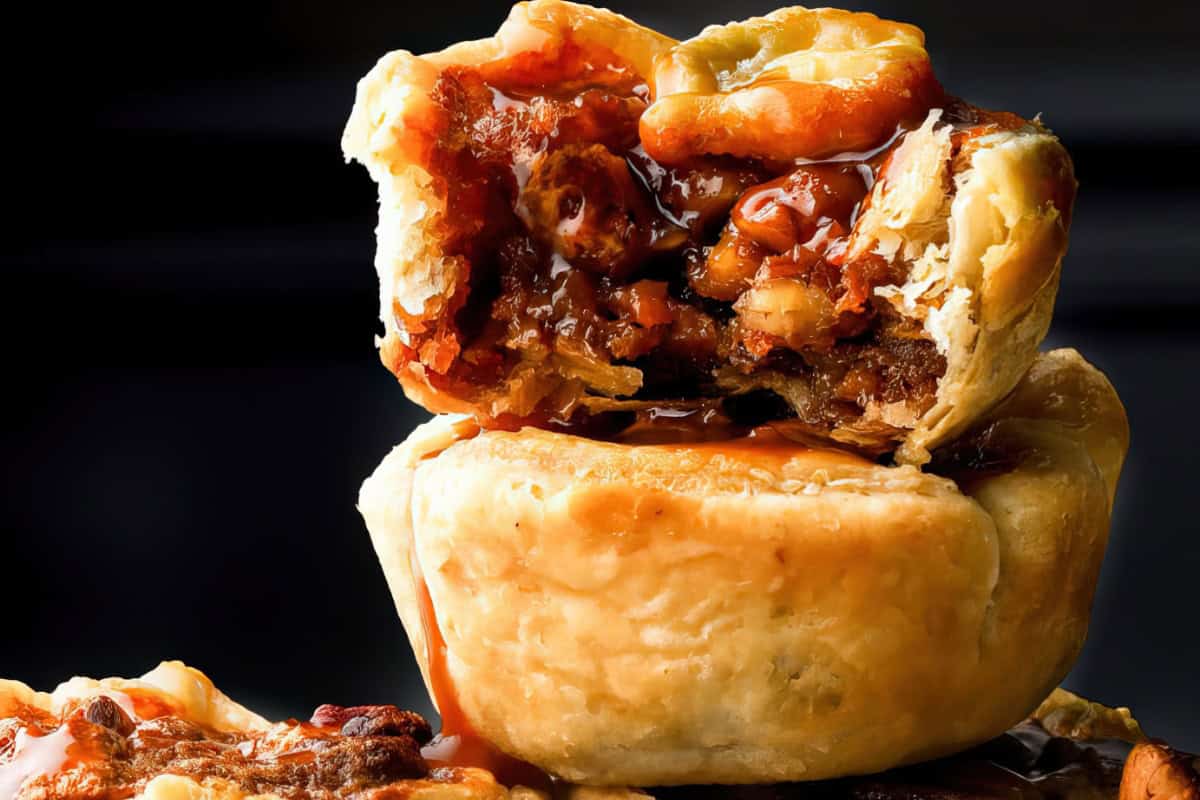 Close-up of a stack of two pecan pies, with the top pie cut open to reveal a gooey, rich filling of pecans and caramelized syrup. The golden, flaky crust contrasts with the rich, dark filling, highlighting the pie's texture.