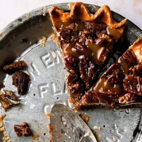A partially eaten pecan pie with caramel topping in a metal tin, with a pie server inside and crumbs scattered around.