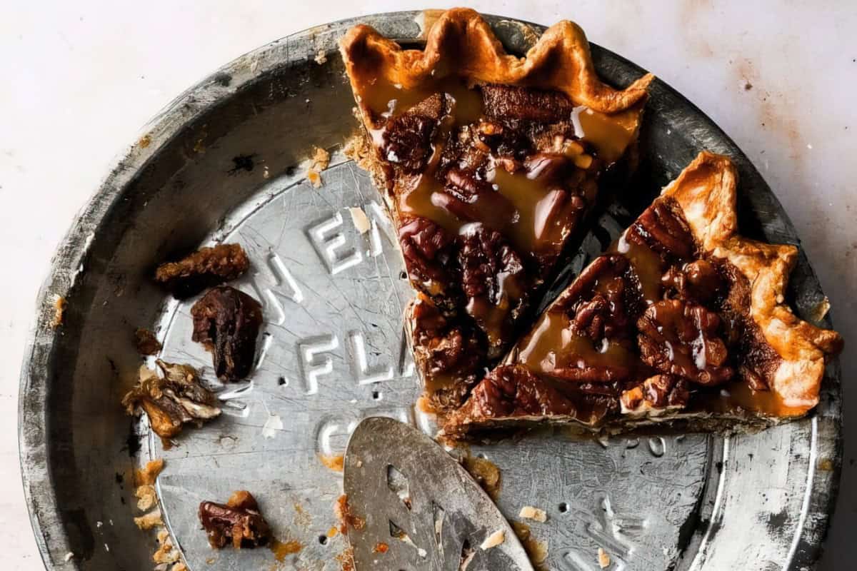 A partially eaten pecan pie in a metal pie dish with a flaky crust. The pie filling is glossy and topped with whole pecans. A pie server rests nearby, with crumbs scattered on the dish. The background is a light-colored surface.