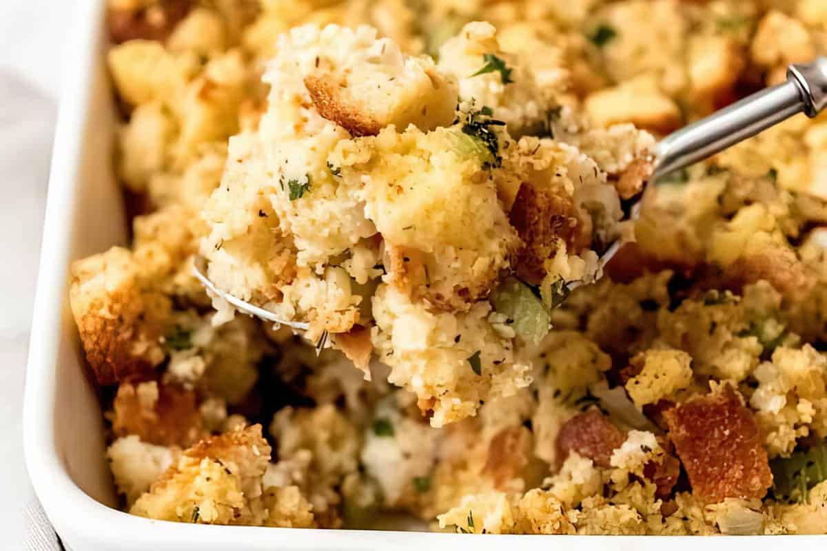 A close-up of a serving spoon lifting a portion of cornbread stuffing from a white baking dish. The stuffing appears golden brown, with visible chunks of bread, herbs, and pieces of celery.