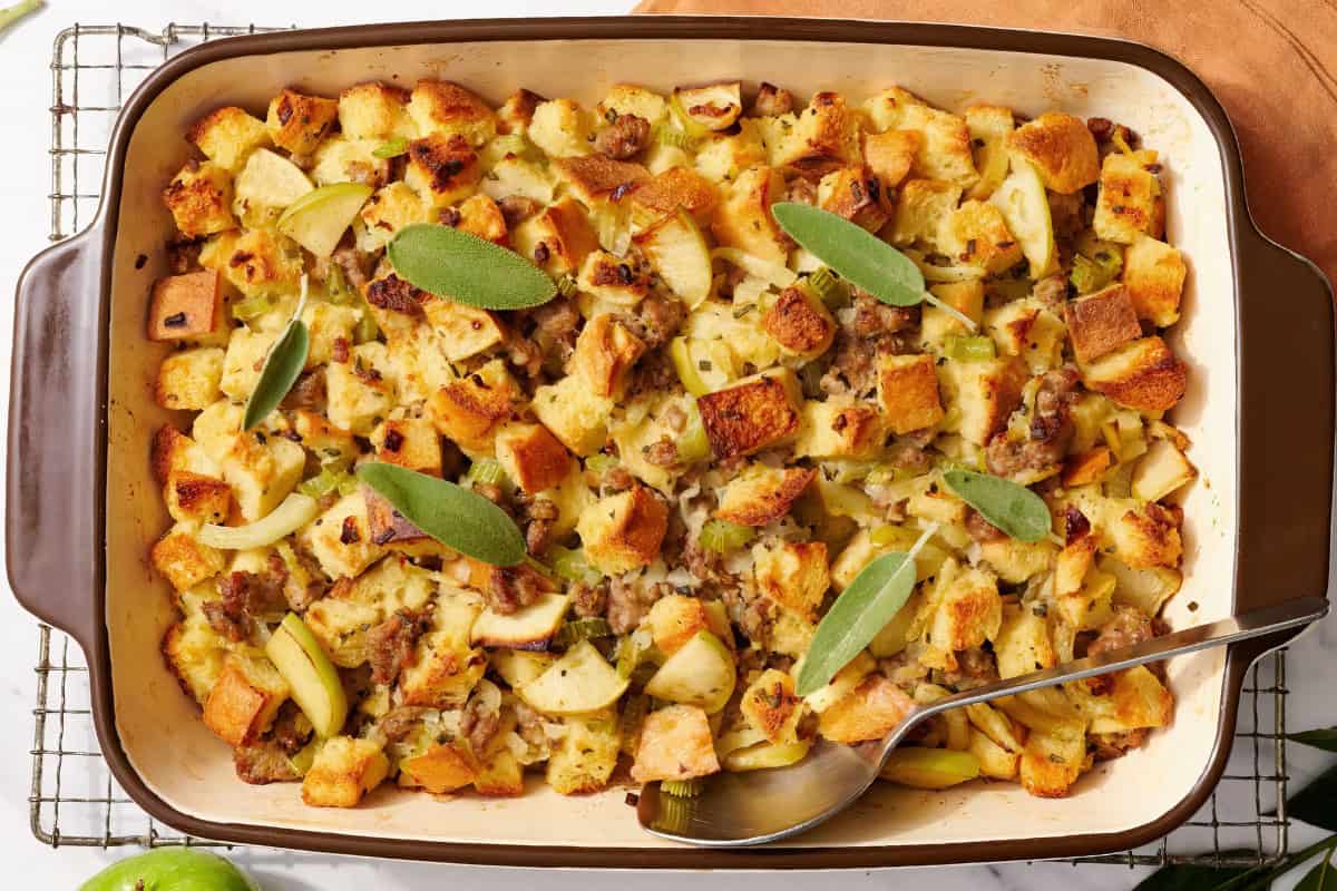 A baked casserole dish filled with golden-brown stuffing, featuring cubes of bread, pieces of sausage, sliced apples, and fresh sage leaves on top. A serving spoon rests in the corner of the dish.