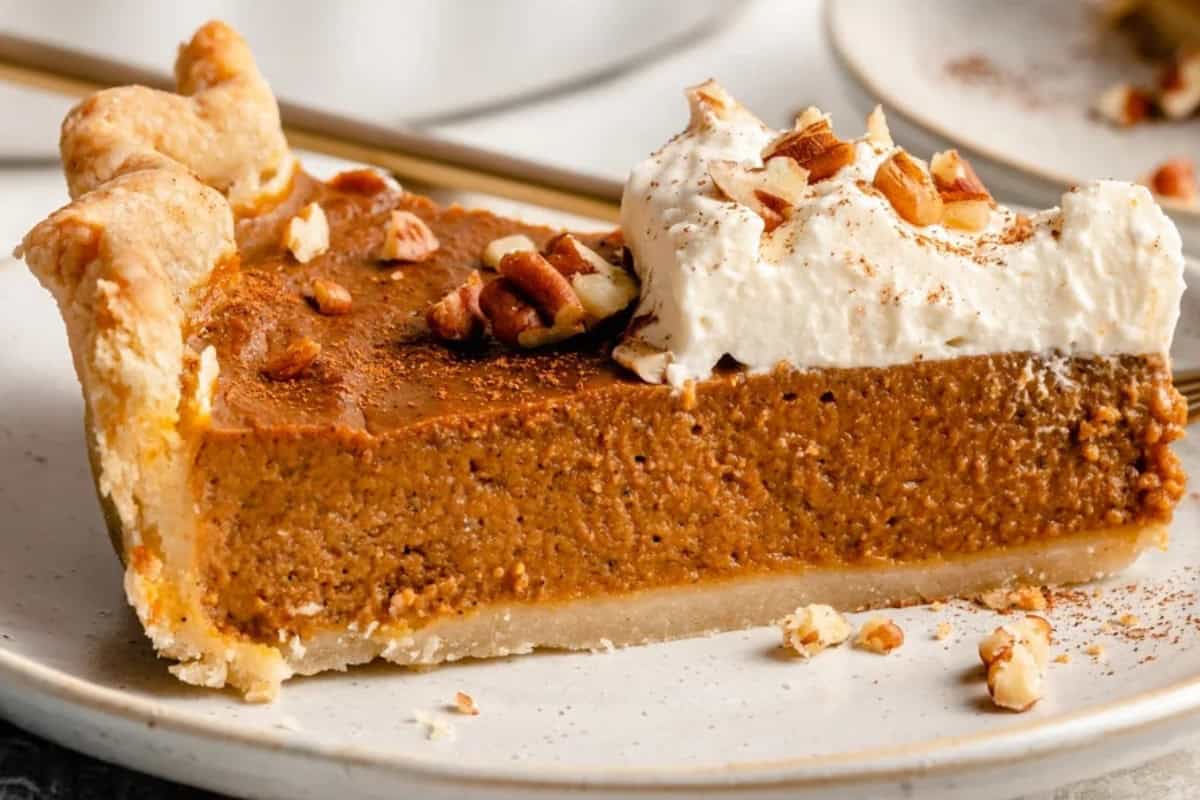 A slice of pumpkin pie topped with whipped cream and chopped pecans on a plate. The pie has a flaky crust and a rich, orange filling.