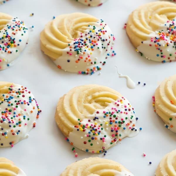 Round swirl cookies partially dipped in white icing with multicolored sprinkles, arranged on a white surface.