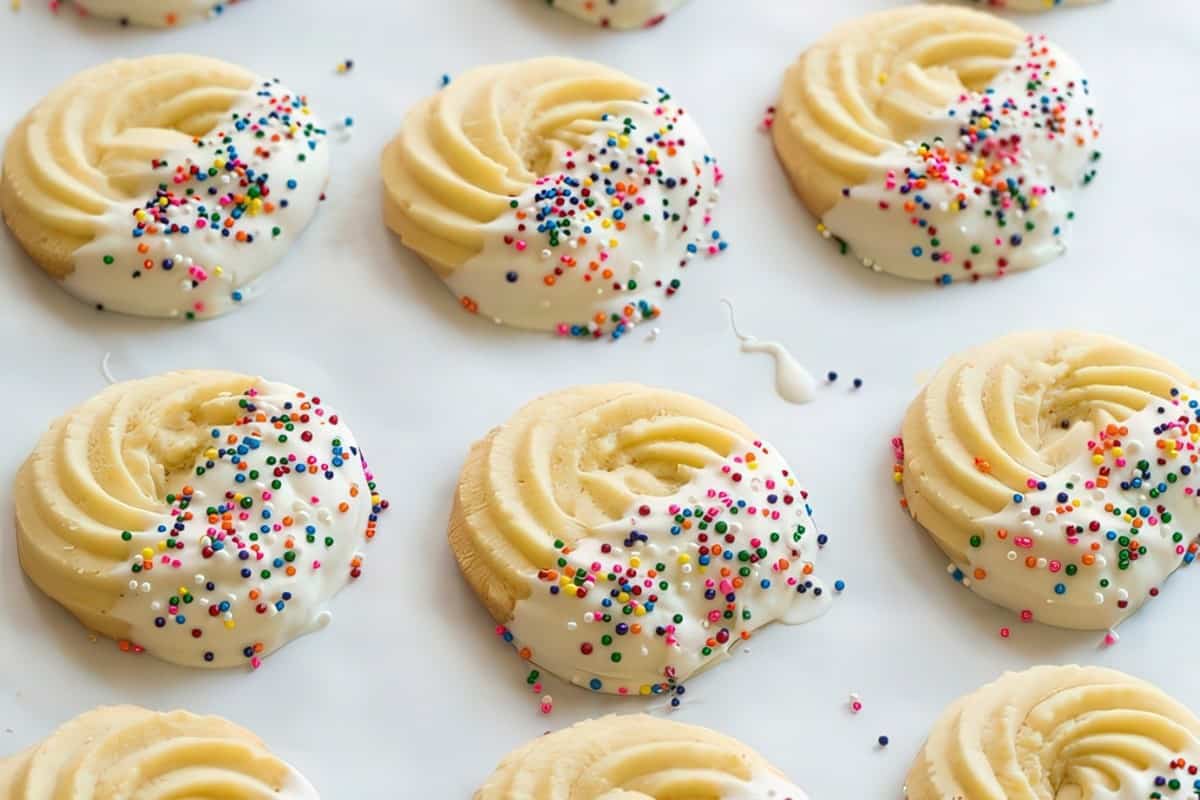 A batch of spiral-shaped cookies partially dipped in white icing, topped with colorful sprinkles. They're arranged on a white surface.