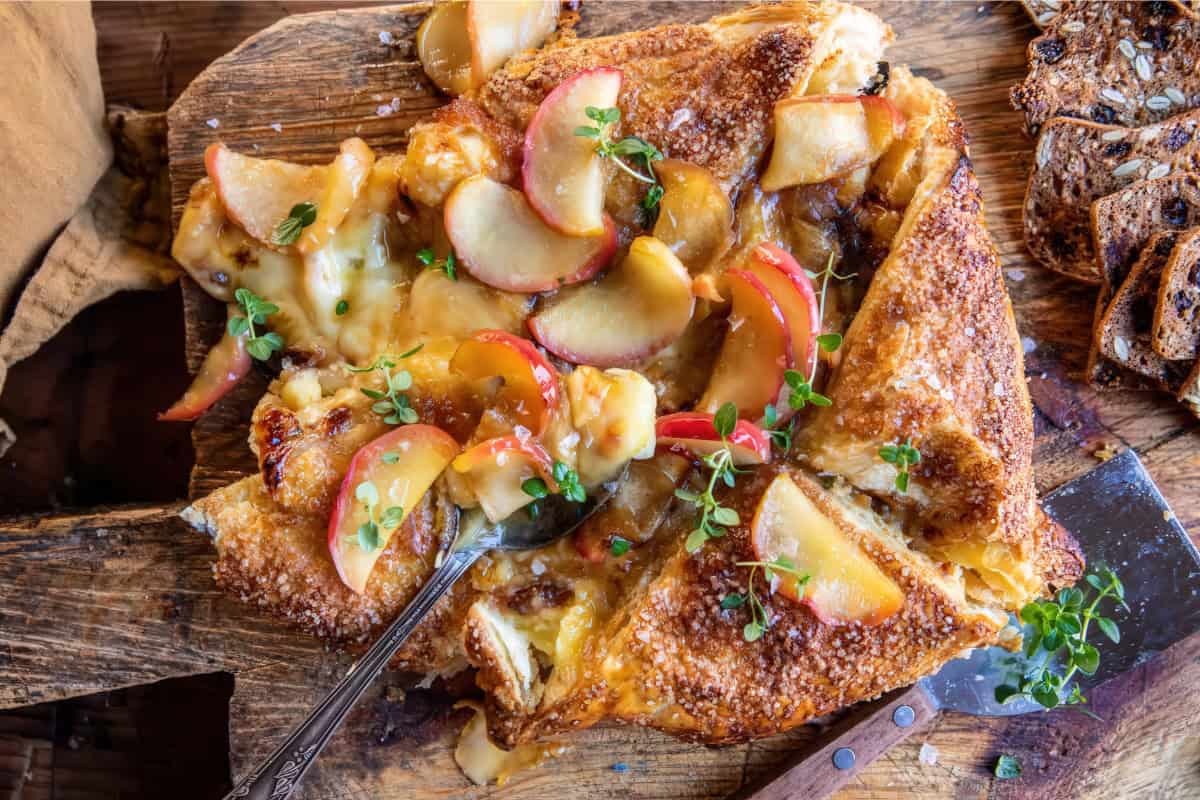 A baked brie topped with sliced apples and herbs on a rustic wooden board. Melted cheese oozes from the golden, crispy pastry. A metal serving spoon rests beside the dish, with slices of grainy bread in the background.