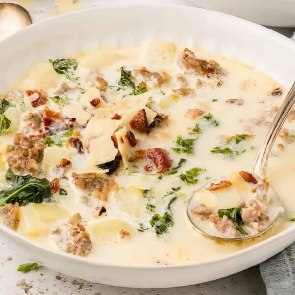 The image features a bowl of creamy soup filled with chopped kale, sausage slices, bacon pieces, potato chunks, and a topping of grated cheese. A spoon rests in the bowl.
