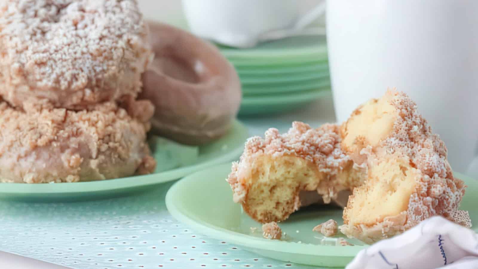 A close-up of a partially eaten crumb cake on a green plate. In the background, there are more pastries stacked on another plate, with a softly focused cup. The scene appears to be a cozy breakfast or dessert setting.
