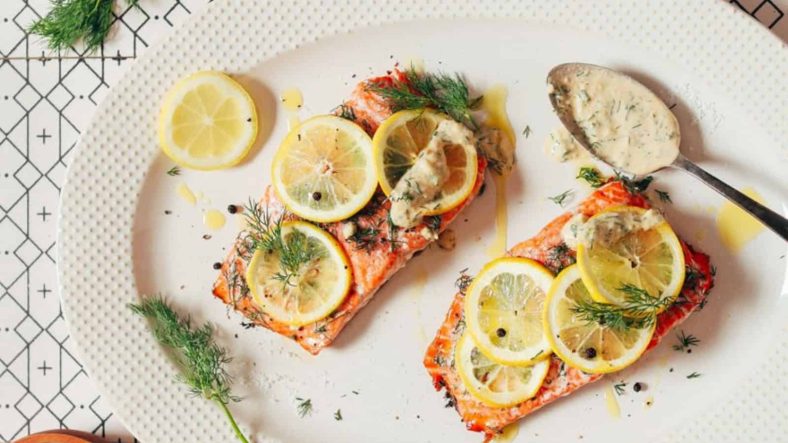 A white plate with two pieces of cooked salmon topped with lemon slices and fresh dill. A spoon with creamy dill sauce rests nearby. The plate sits on a patterned tablecloth.
