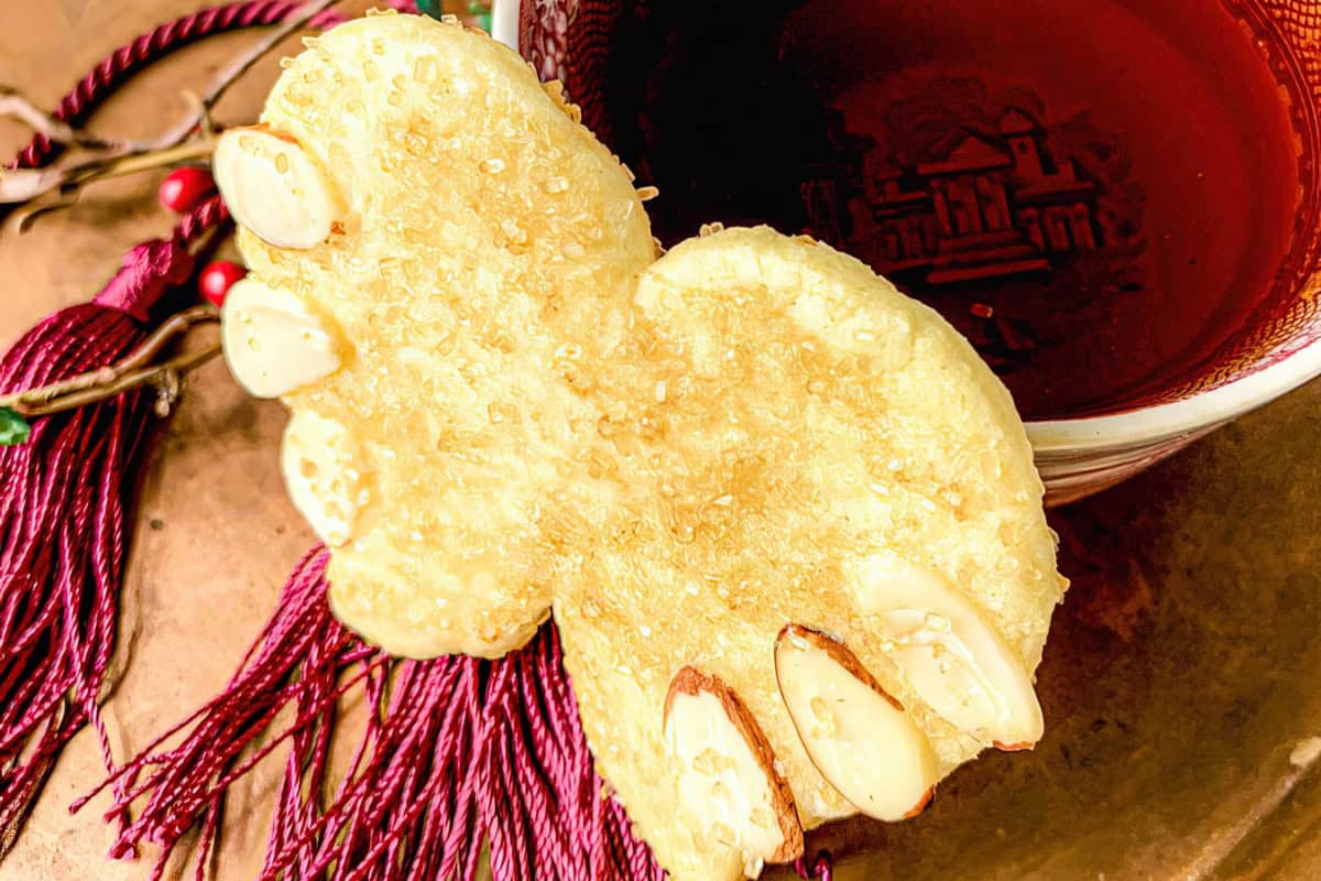 An angel wing-shaped cookie with almond slices rests on a bronze tray. Nearby, there is a red decorative cloth and a golden ornament.