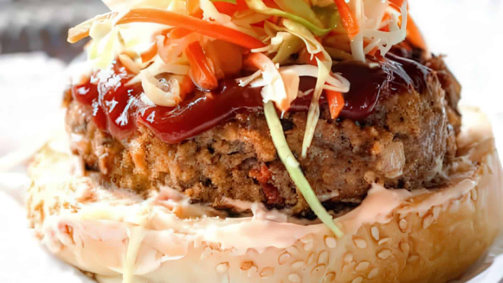 Close-up of a burger with a sesame seed bun. The patty is topped with barbecue sauce and a heap of coleslaw consisting of shredded cabbage and carrots. The bottom bun is spread with a creamy dressing.
