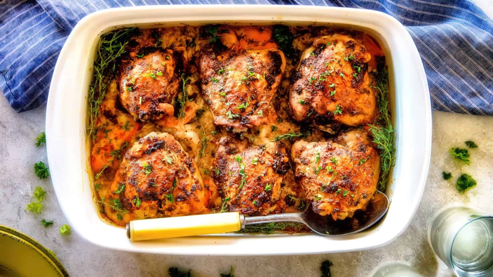 Baked chicken thighs in a white casserole dish, garnished with fresh herbs. A spoon rests at the side. A striped blue cloth and a green plate are nearby on a light-colored surface.