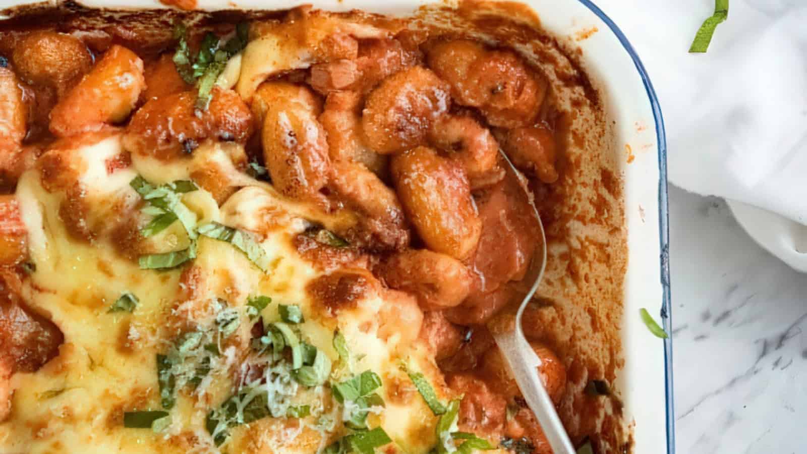 A close-up of a baked gnocchi dish in a pan, topped with melted cheese, tomato sauce, and fresh basil. A serving spoon rests in the dish, and the background shows a white marble surface.