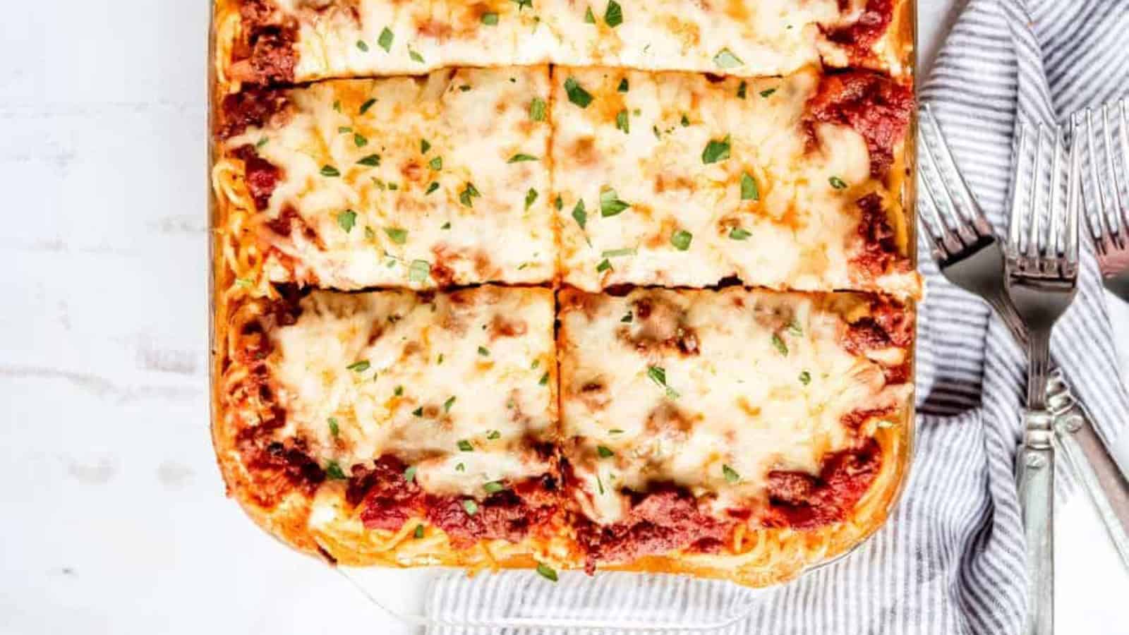 A freshly baked lasagna with a golden, bubbly cheese topping and a rich tomato meat sauce. The dish is cut into six rectangular pieces, garnished with fresh herbs. Two forks rest on a striped cloth beside the dish.