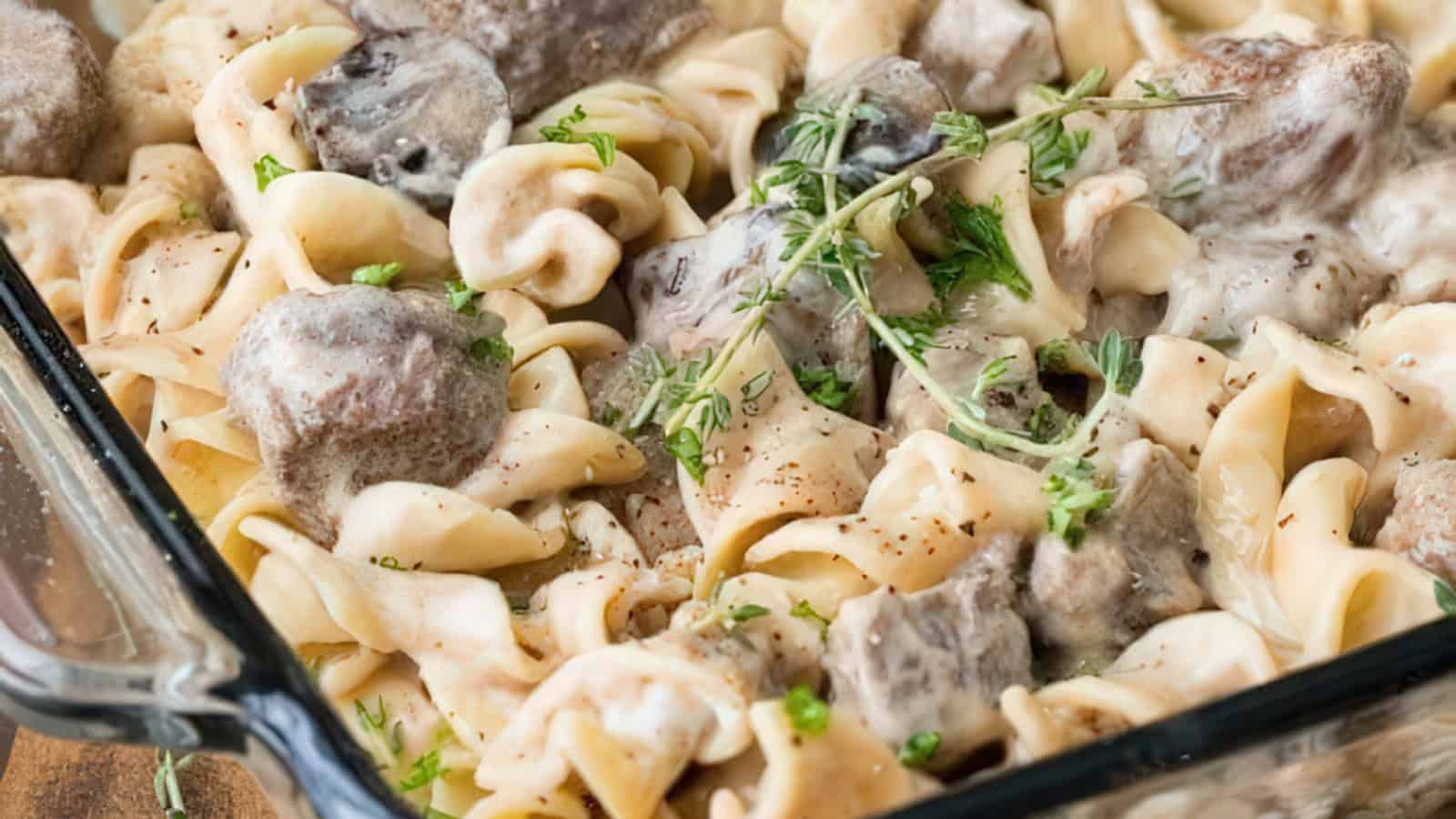 A close-up of a delicious beef stroganoff dish with tender beef chunks and wide egg noodles coated in creamy sauce, garnished with fresh herbs. The meal is served in a glass baking dish.