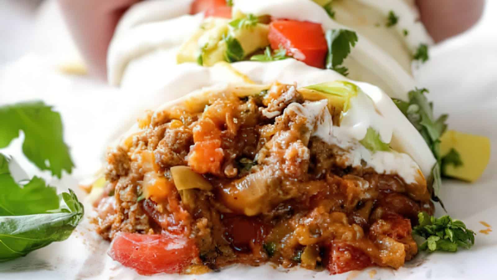 Close-up of a burrito with a bite taken out, revealing seasoned ground meat, melted cheese, diced tomatoes, and fresh cilantro. The burrito is topped with sour cream and surrounded by a few leaves of cilantro on a paper-lined surface.