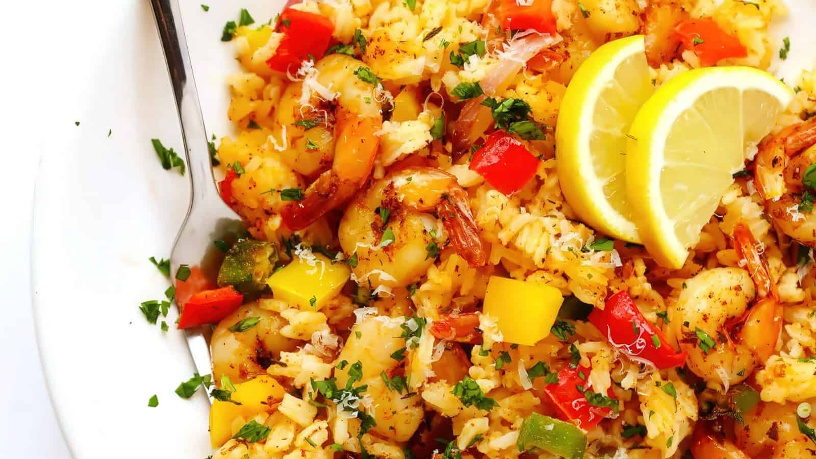 A plate of shrimp and rice stir-fry, garnished with chopped herbs. The dish includes red and yellow bell peppers, topped with grated cheese and lemon slices. A fork rests on the side of the plate.