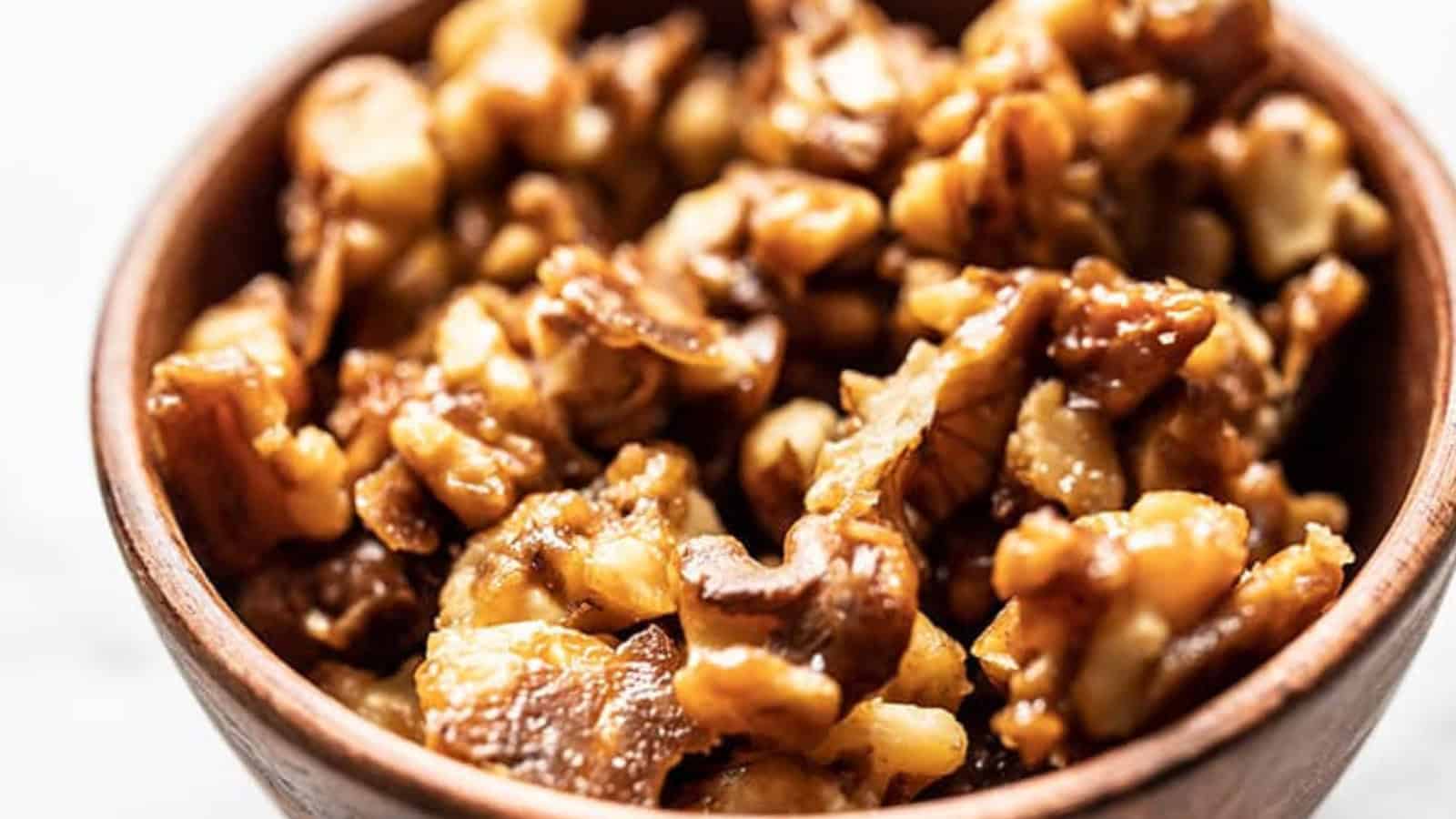 A close-up image of a bowl filled with walnuts. The nuts have a rich, brown color and a slightly glossy texture, suggesting they are fresh or possibly roasted. The bowl is made of wood and sits on a light-colored surface.