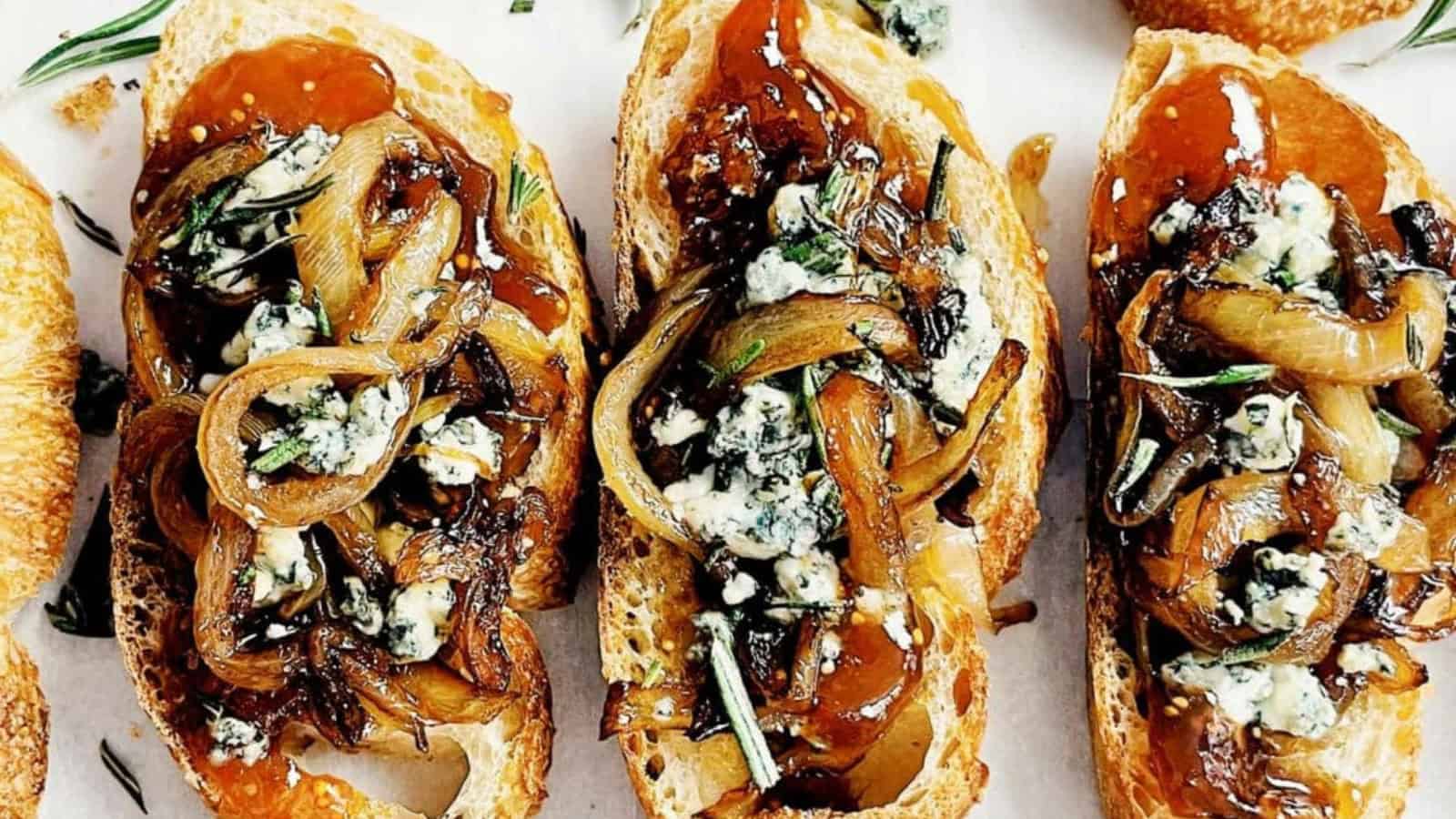 Toasted baguette slices topped with caramelized onions, blue cheese crumbles, and a drizzle of glaze, garnished with fresh rosemary.