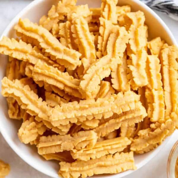 A bowl filled with golden, crinkle-cut cookies sits on a white surface. Some cookies are scattered around the bowl. A patterned cloth is partially visible at the top right corner.