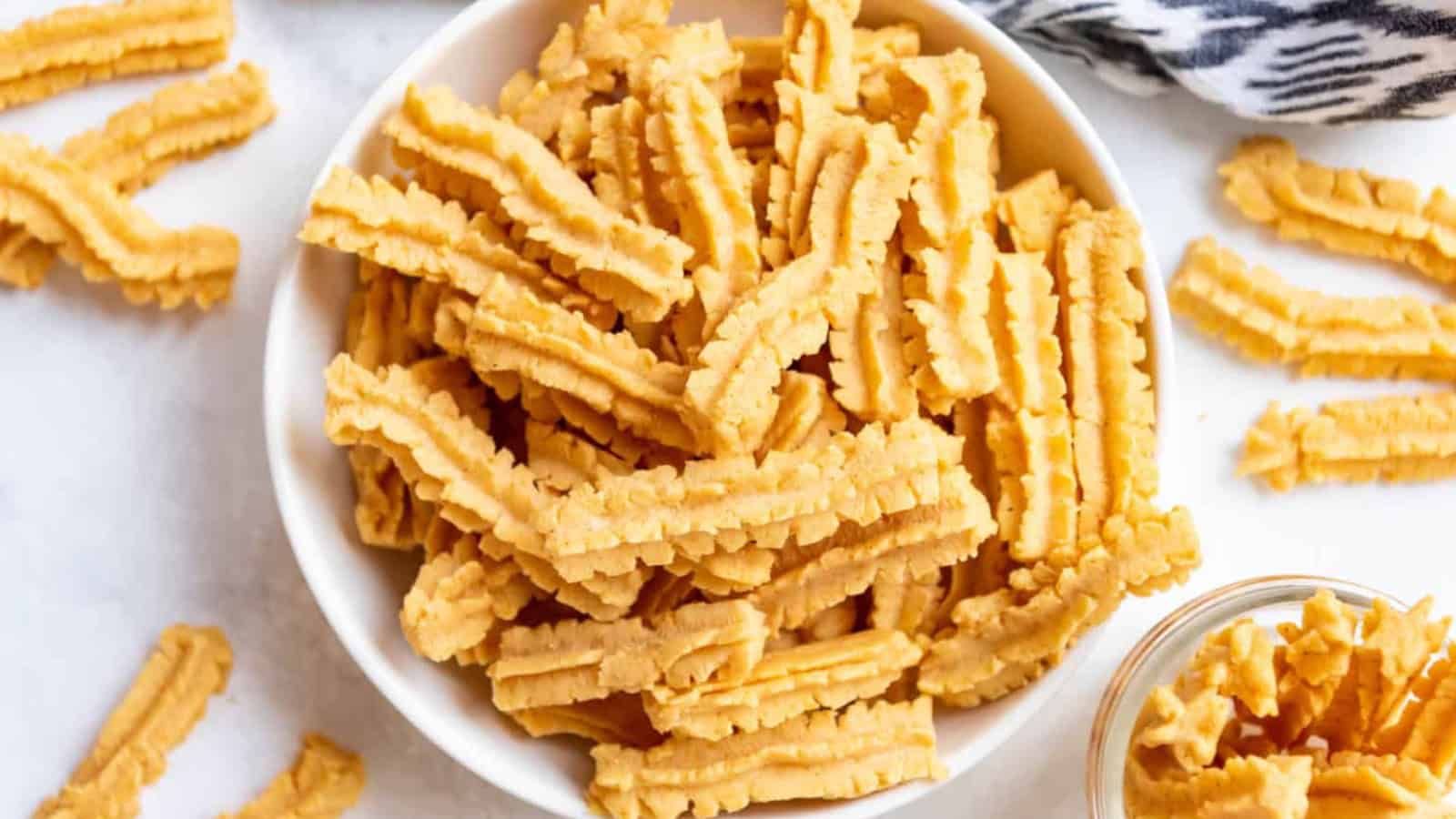 A bowl filled with golden, crinkle-cut cookies sits on a white surface. Some cookies are scattered around the bowl. A patterned cloth is partially visible at the top right corner.