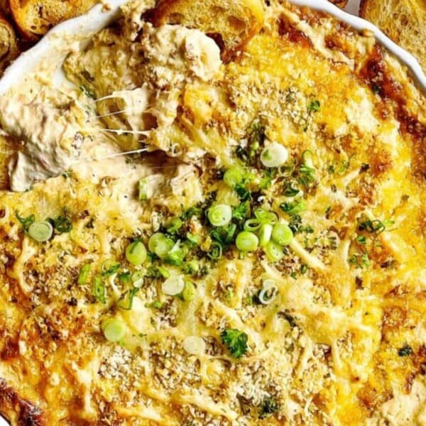 A hand dips toasted bread into baked cheese and artichoke dip with green onions on a white plate surrounded by extra toast.