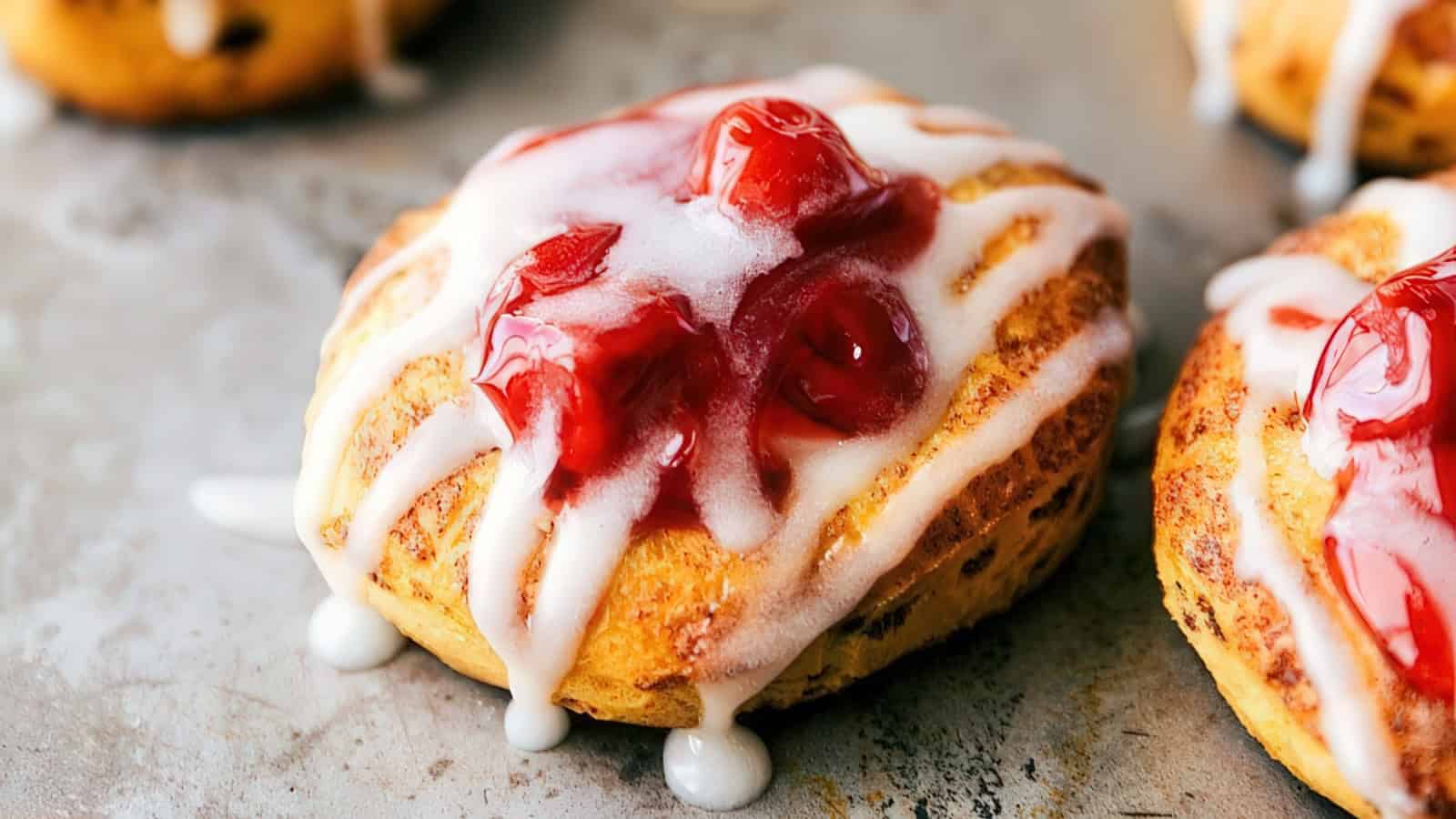 A freshly baked pastry topped with red cherry filling and drizzled with white icing rests on a baking sheet, with another similar pastry partially visible in the background.