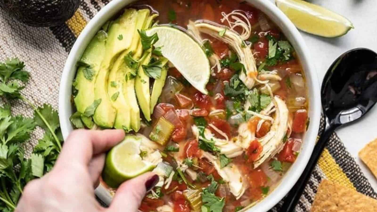 A bowl of chicken tortilla soup topped with sliced avocado, lime wedges, and cilantro. A hand squeezes a lime over the soup. A black spoon and tortilla chips are on the side, with cilantro leaves scattered around.